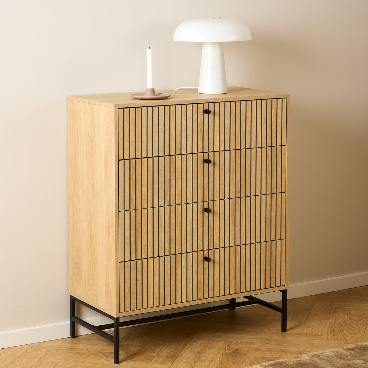 Die Kommode „Bambouz“ (81 x 100) vor einer beigen Wand mit einer weißen Lampe und einer Kerze auf einem Parkettboden im Fischgrätenmuster – eine Kommode für das Schlafzimmer