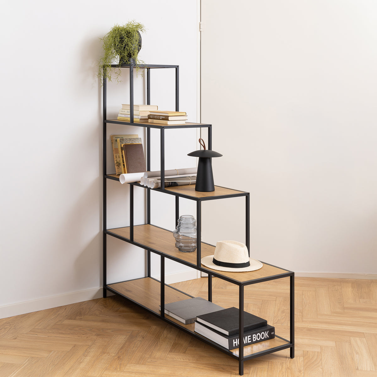 Das Regal „Skeleto“ 135 x 150 in der Farbe „Wilde Eiche“ in der Ecke des Zimmers, mit Büchern, einer Lampe und einem Hut – ein stufenförmiges Regal