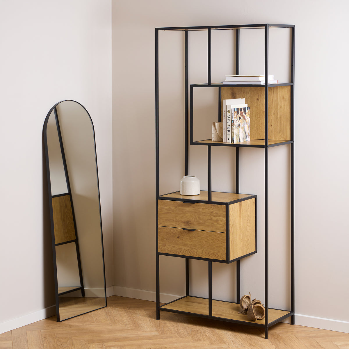 Das Regal „Skeleto“ (77 x 185) in der Ausführung „Wilde Eiche“ in einem hellen Innenraum neben einem bogenförmigen Spiegel, mit Büchern und einer Vase auf den Regalböden
