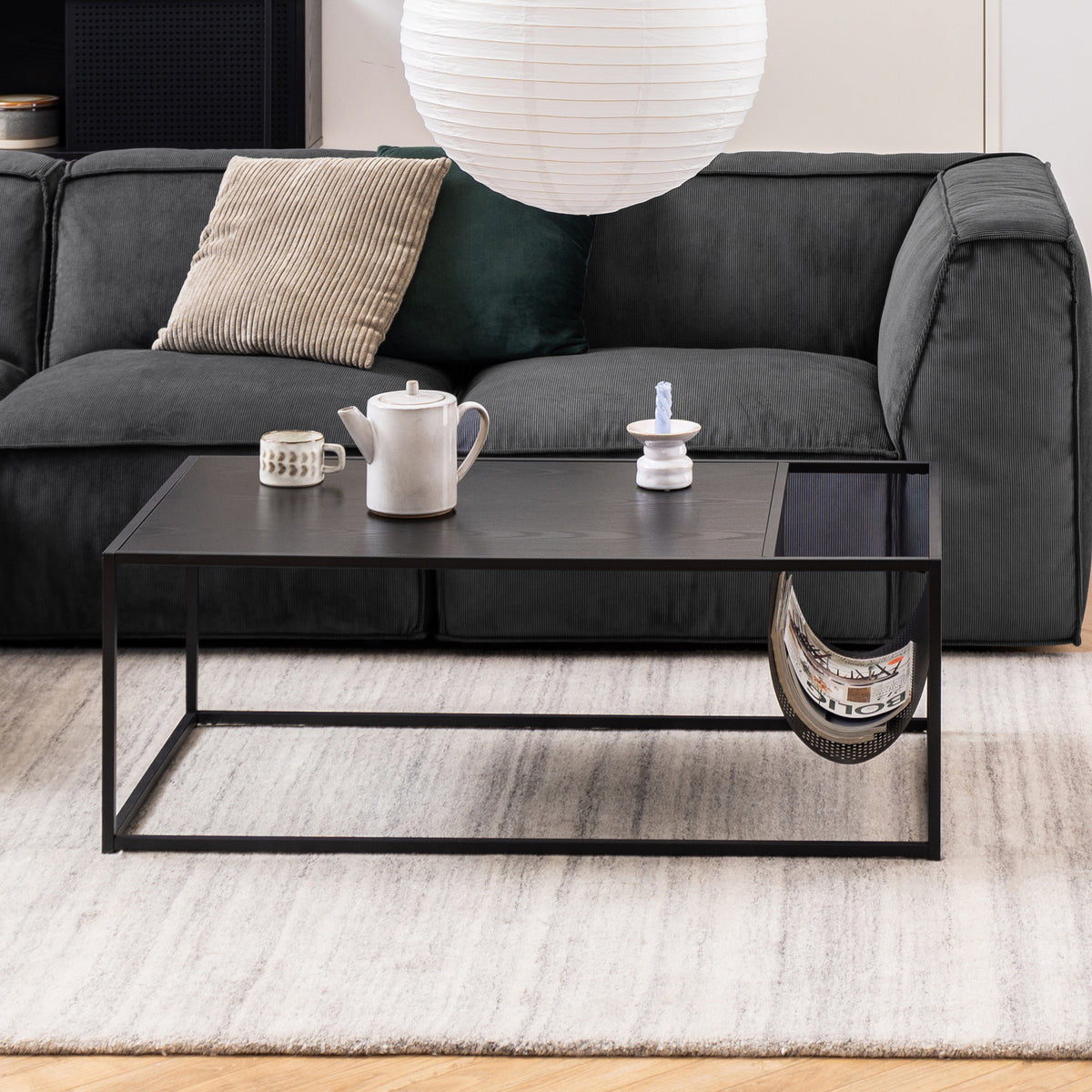 Der Couchtisch „Skeleto“ (110 x 60) vor einem graphitfarbenen Sofa auf einem hellen Teppich, mit einer Kanne und Zeitschriften in einer Metalltasche