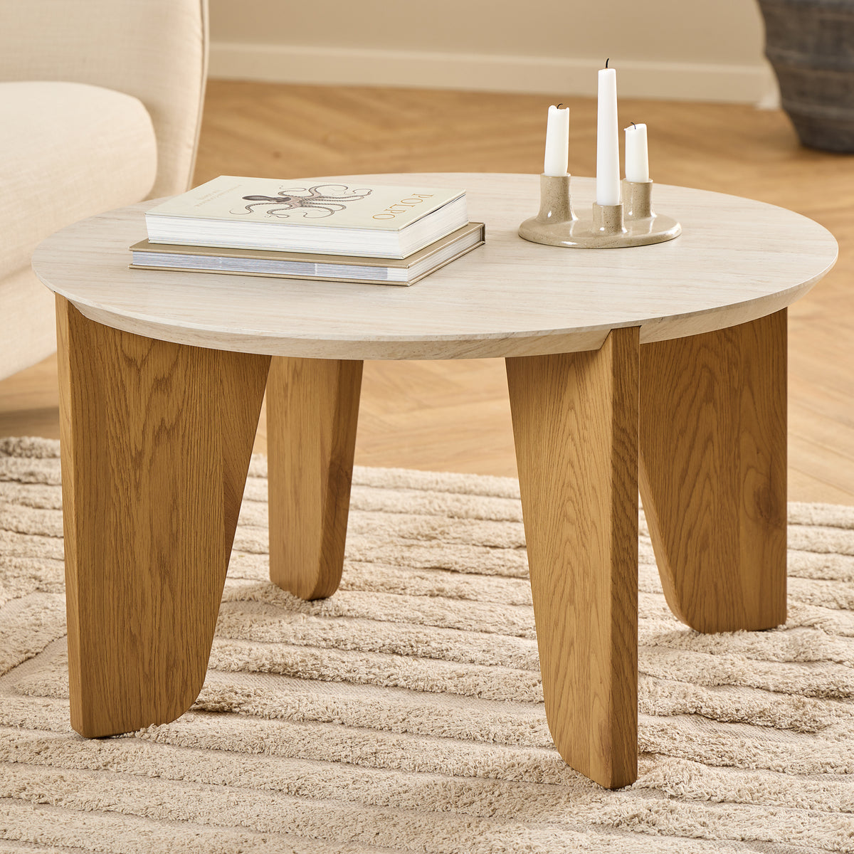 Der Couchtisch „Tendra“ (Ø 80 × 45) auf einem beigen Teppich neben einem hellen Sessel, mit Büchern und einem Kerzenständer – ein Tisch für das Wohnzimmer
